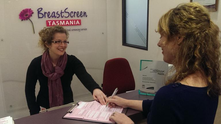 A breast screen tasmania worker handing a woman a sign in sheet at the reception area.